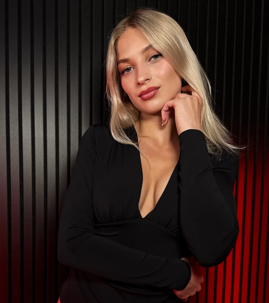 FairyFox blond im schwarzen Longsleeve-Kleid mit tiefem Ausschnitt vor rot beleuchteter Lamellenwand